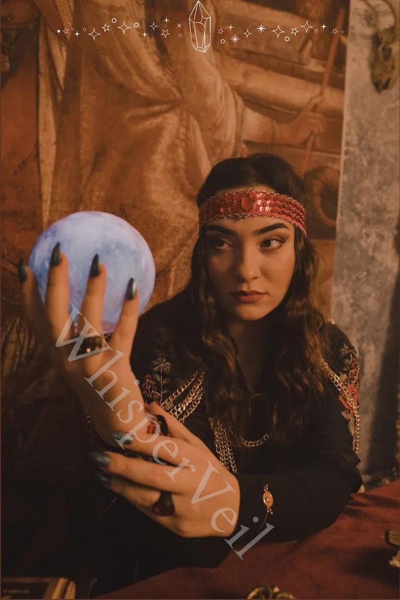 May include: A woman wearing a red sequined headband and a black top with gold chain accents holds a glowing blue orb. The image has a mystical theme, with the text "WhisperVeil" partially visible.