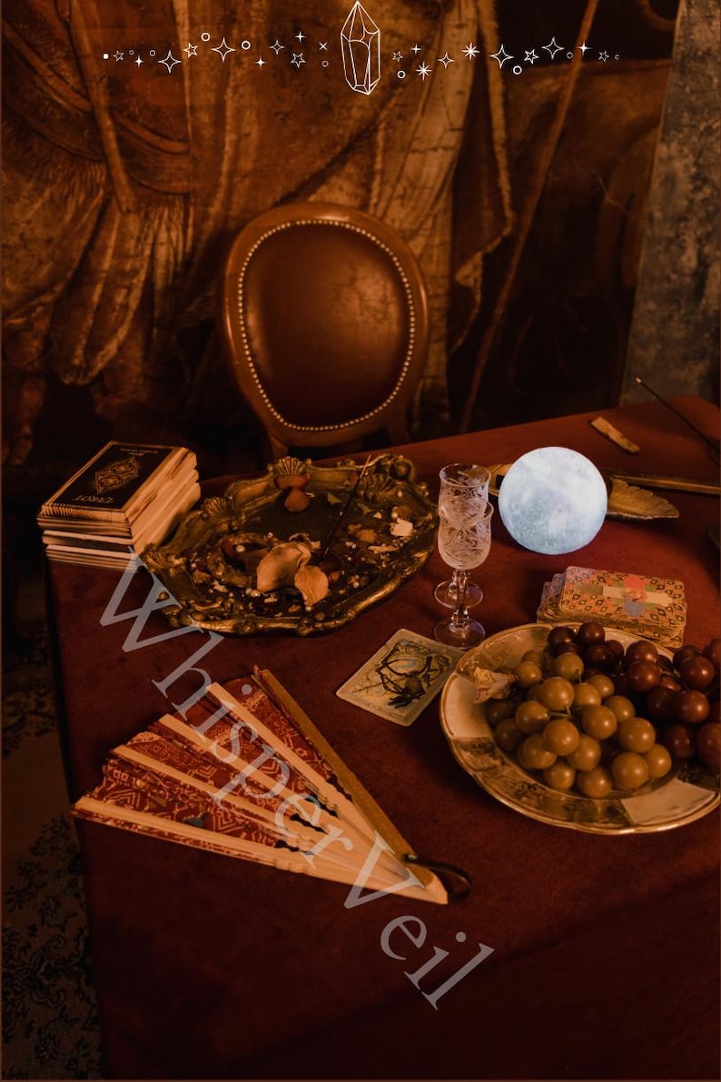 Puede incluir: Un bodeg&oacute;n con una mesa roja, un abanico decorativo, una pila de libros, una bandeja decorativa, una copa, una luz con forma de luna, un plato de uvas y cartas del tarot. El texto "Whisper Veil" es visible.