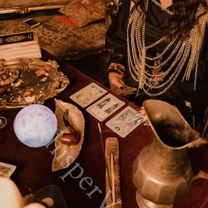 Puede incluir: Una escena m&iacute;stica con cartas del tarot, un orbe brillante y un porta incienso con estrellas. Una jarra de metal y una bandeja decorativa est&aacute;n sobre una superficie de terciopelo rojo. El entorno sugiere un tema espiritual o de adivinaci&oacute;n.