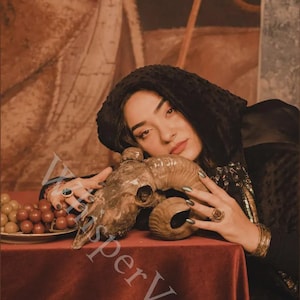 May include: A still life featuring a ram skull, grapes, and a red tablecloth. The skull is brown with curved horns. Grapes are on a white plate. The background is a sepia-toned painting.