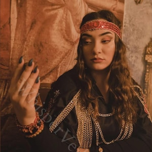 May include: A woman wearing a red sequined headband and a black top adorned with gold chains. The image has a vintage aesthetic, with a warm color palette and a decorative background. The woman is also wearing a red bracelet.