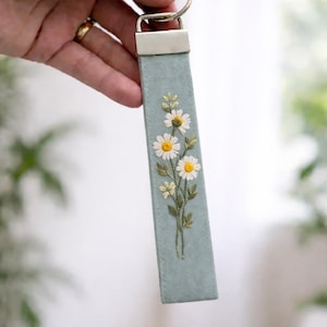 May include: A rectangular fabric keychain in a soft sage green color featuring detailed embroidery of three white daisies with yellow centers and green stems. The fabric strip is attached to a silver-toned metal hardware clamp and a circular ring.