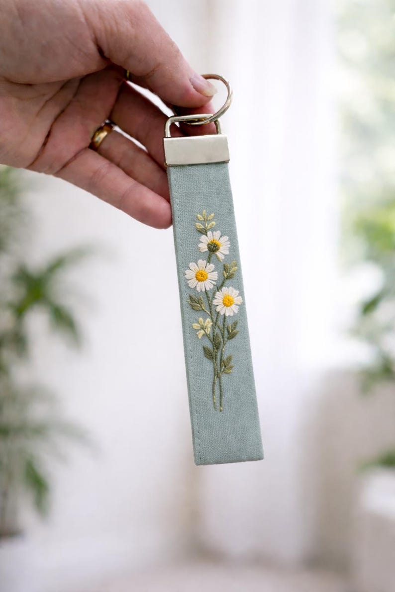May include: A rectangular fabric keychain in a soft sage green color featuring detailed embroidery of three white daisies with yellow centers and green stems. The fabric strip is attached to a silver-toned metal hardware clamp and a circular ring.