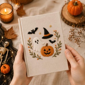 Peut inclure: Un livre à couverture rigide beige orné de motifs d'Halloween brodés sur la couverture, incluant un fantôme blanc, une citrouille orange, un chapeau de sorcière noir, deux chauves-souris noires, des étoiles jaunes et des tiges vertes.