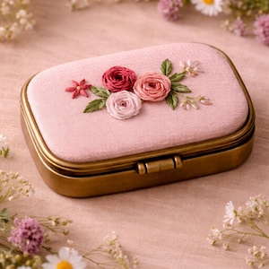 Puede incluir: Una caja de joyería rectangular rosa con un marco y cierre color latón. La tapa está adornada con rosas bordadas en tonos rosa y rojo, junto con hojas verdes. La caja está rodeada de flores.