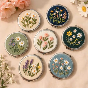 May include: A collection of seven round, silver-framed compact mirrors. Each mirror features a different embroidered floral design, including daisies, roses, and lavender, on various colored backgrounds such as cream, navy, and green. The mirrors are arranged on a light-colored surface.