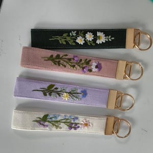 May include: Four fabric wristlet keychains with gold-tone metal hardware. Each strap features unique floral embroidery: white daisies on black, purple flowers on dusty pink, blue and yellow blossoms on lavender, and mixed pastel flowers on cream.