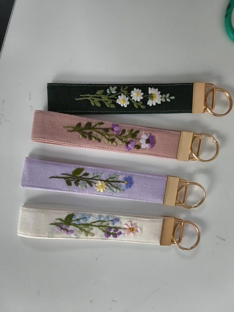 May include: Four fabric wristlet keychains with gold-tone metal hardware. Each strap features unique floral embroidery: white daisies on black, purple flowers on dusty pink, blue and yellow blossoms on lavender, and mixed pastel flowers on cream.