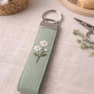 May include: A sage green fabric keychain featuring an embroidered design of two white daisies with yellow centers and green stems. The rectangular fabric strap is attached to a silver-toned metal split ring by a matching silver-toned metal clamp.