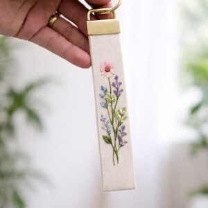 May include: A rectangular fabric keychain featuring an off-white base with an embroidered floral design. The embroidery depicts a single pink flower at the top, surrounded by blue and purple sprigs and green leaves. It has a gold-toned metal clasp.