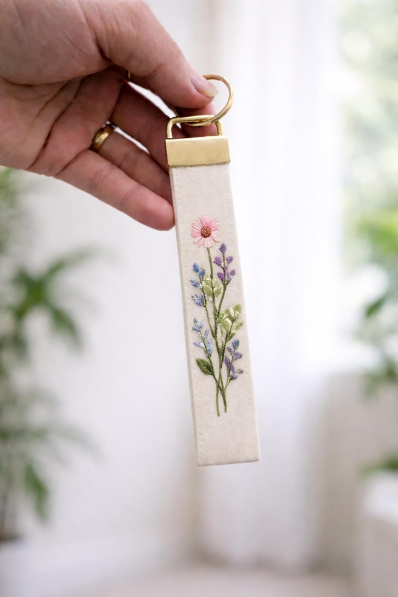 May include: A rectangular fabric keychain featuring an off-white base with an embroidered floral design. The embroidery depicts a single pink flower at the top, surrounded by blue and purple sprigs and green leaves. It has a gold-toned metal clasp.