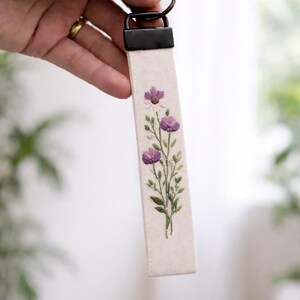 May include: A rectangular fabric keychain featuring an off-white canvas base with an embroidered floral design. The embroidery depicts three purple flowers with green stems and leaves. A black metal hardware clamp secures the fabric to a metal ring.