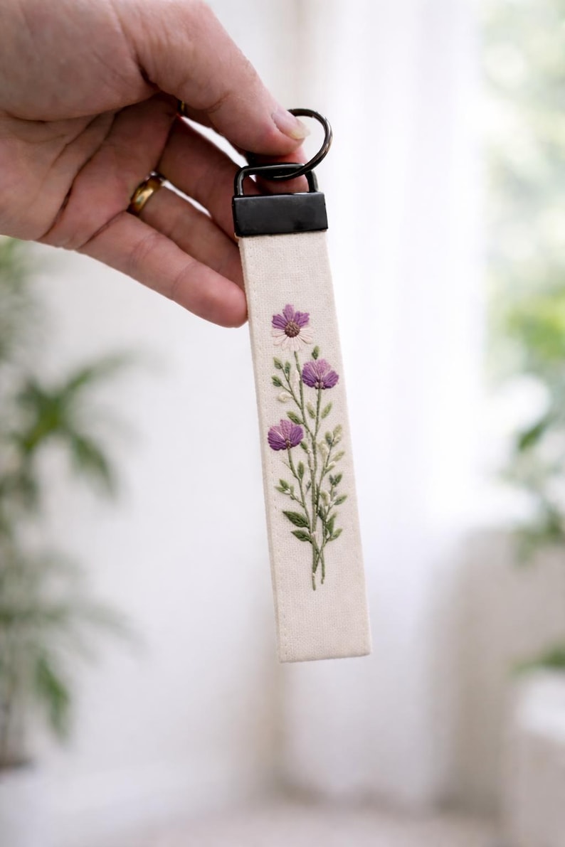 May include: A rectangular fabric keychain featuring an off-white canvas base with an embroidered floral design. The embroidery depicts three purple flowers with green stems and leaves. A black metal hardware clamp secures the fabric to a metal ring.