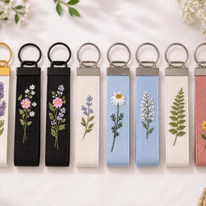 Könnte beinhalten: Acht rechteckige Schlüsselanhänger aus Stoff in einer Reihe, jeder mit einzigartiger Blumen- oder Pflanzenstickerei. Die Designs umfassen Lavendel, Gänseblümchen, Wildblumen und Farne auf verschiedenfarbigen Stoffbändern mit Metallringen.