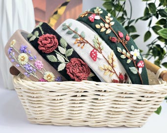 May include: A collection of five embroidered headbands in a woven basket. The headbands feature floral and botanical designs in various colors, including purple, red, and green. The headbands are displayed in a light-colored woven basket.