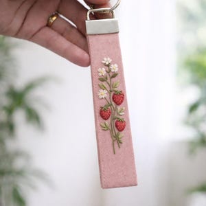 May include: A pink fabric keychain featuring an embroidered design of three red strawberries with green leaves and small white flowers. The fabric strap is attached to a silver-toned metal hardware clamp and a circular split ring for keys.