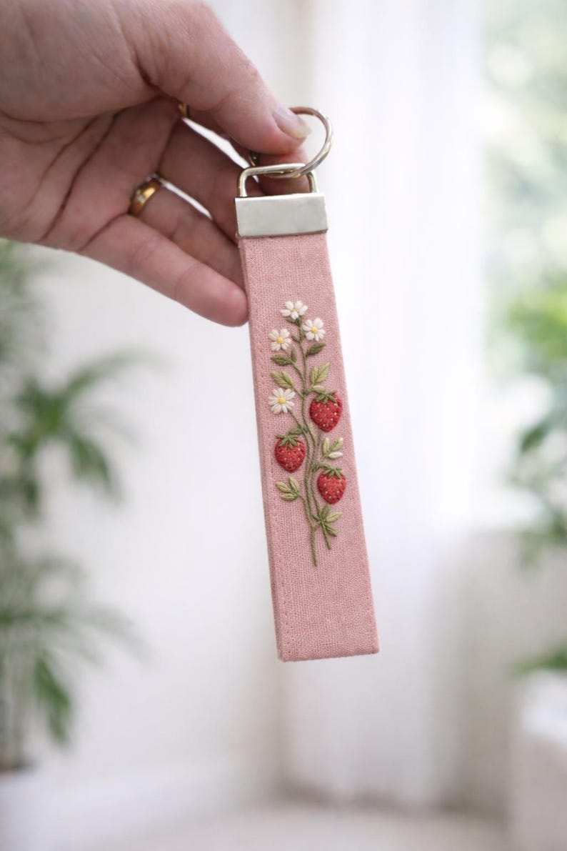 May include: A pink fabric keychain featuring an embroidered design of three red strawberries with green leaves and small white flowers. The fabric strap is attached to a silver-toned metal hardware clamp and a circular split ring for keys.