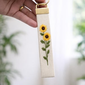 May include: A rectangular off-white fabric keychain featuring an embroidered design of two yellow sunflowers with brown centers and green leaves on a stem. The fabric strip is attached to a gold-toned metal hardware clasp and a circular ring.