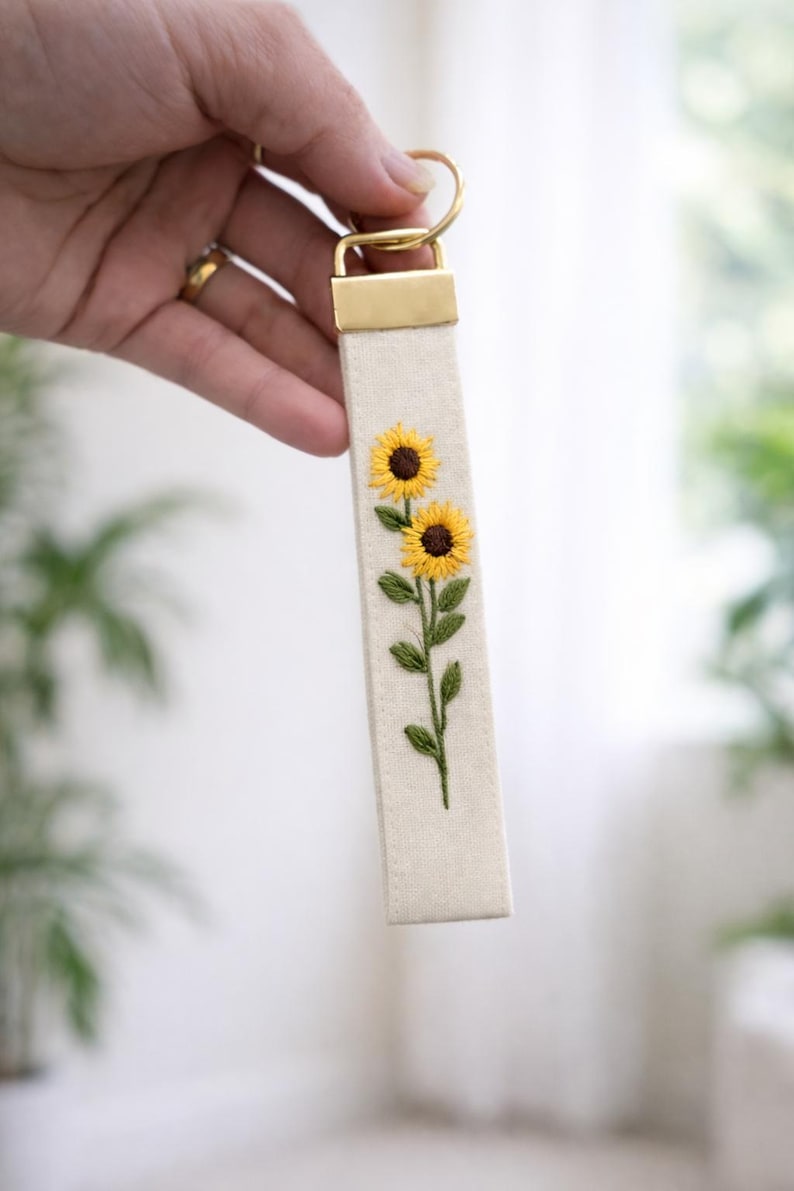 May include: A rectangular off-white fabric keychain featuring an embroidered design of two yellow sunflowers with brown centers and green leaves on a stem. The fabric strip is attached to a gold-toned metal hardware clasp and a circular ring.