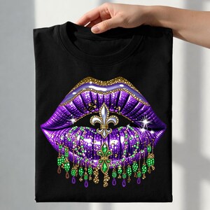 May include: Black t-shirt featuring a graphic of purple lips with gold glitter accents. A fleur-de-lis and dangling green and gold beaded embellishments are centered on the lips. The design is a vibrant and eye-catching statement.