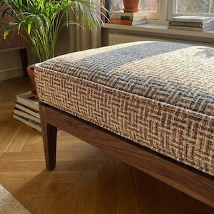 Könnte beinhalten: Ein rechteckiger Hocker mit einem dunklen Holzrahmen und einem gemusterten Kissen. Das Kissen hat ein gewebtes Design in Braun-, Beige- und Grautönen. Der Hocker steht auf einem Parkettboden, mit einer Pflanze und Büchern im Hintergrund.