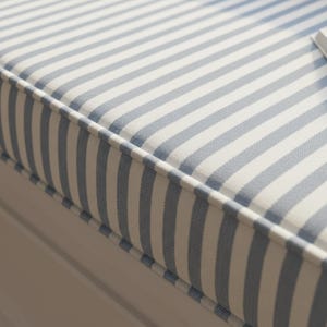 May include: Close-up of a striped cushion with a white and light blue pattern. The cushion has a white border and is likely part of a bench or window seat. The fabric has a textured appearance.