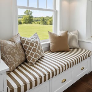 Könnte beinhalten: Fensterbank mit gestreiftem Kissen und mehreren Zierkissen. Die Kissen haben verschiedene Muster, darunter Blumen- und geometrische Designs. Der Sitz ist weiß mit Messing-Schubladengriffen. Das Fenster bietet einen Blick auf eine grüne Wiese und Bäume.