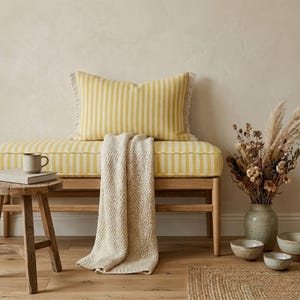 Puede incluir: Un banco a rayas amarillas y blancas con un cojín a juego y una manta color crema. Un pequeño taburete de madera sostiene una taza y un libro. Un jarrón de flores secas y cuencos pequeños completan el interior de tonos neutros.