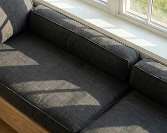 May include: A dark gray upholstered window seat with a light wood frame. The seat has a rectangular shape with a backrest and two bolster pillows. The window is white and has a view of green grass.