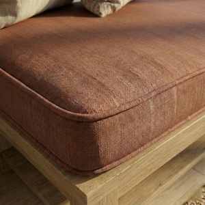 Può includere: Primo piano di una sezione di una panca in legno con un cuscino color ruggine. Il cuscino ha un tessuto strutturato e un cordoncino a contrasto. Il telaio della panca è in legno chiaro.