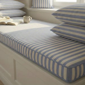 May include: A window seat cushion and pillows with a blue and white striped pattern. A book and a mug are on the cushion. The pillows are stacked on the left and right side of the window seat.