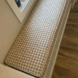 May include: A rectangular bench cushion with a houndstooth pattern in shades of beige and brown. The cushion is bordered with a light beige piping and is set into a white frame, suitable for a window seat or built-in bench.