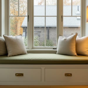 May include: A window seat with a green cushion and four beige pillows. The window frame is white, and the seat has two drawers with brass handles. The background shows a house and trees.