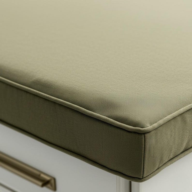 May include: Close-up of a rectangular olive green cushion with a piped edge. The cushion is made of a textured fabric and appears to be part of a bench or seat. The cushion is resting on a white surface.