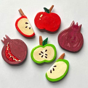 May include: Hand-painted wooden magnets shaped like fruits. The set includes a whole red apple, a green apple half, two pomegranate shapes, and two apple slices. The magnets are painted in bright colours with a white background.