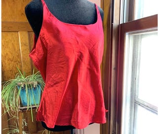 May include: A bright red camisole top with thin straps and a square neckline. The top is displayed on a black mannequin. The fabric appears to be slightly textured. The background includes a wooden door, a plant, and a window.