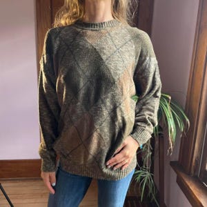May include: A long-sleeved sweater in shades of olive green, brown, and tan, featuring a classic argyle pattern. The sweater has a crew neck and ribbed cuffs and hem. The fabric appears to be a soft knit, suitable for cooler weather.