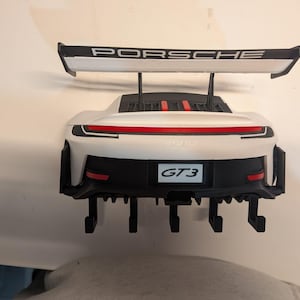 Puede incluir: Modelo Porsche GT3 blanco y negro con alerón trasero. La palabra "PORSCHE" está impresa en el alerón. El modelo tiene un parachoques trasero negro con el texto "GT3".