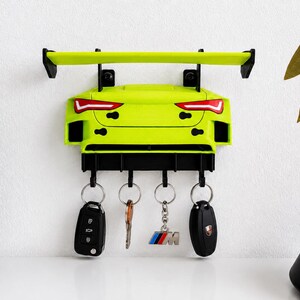 Puede incluir: Un soporte para llaves de pared de color verde lima con forma de parte trasera de coche deportivo, que presenta un alerón negro prominente, luces traseras integradas y cuatro ganchos negros. Cuatro juegos de llaves cuelgan de los ganchos.