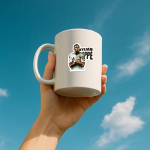 May include: A white ceramic mug featuring a sticker of Kylian Mbappe with the text "KYLIAN MBAPPE". The mug is held against a blue sky background.