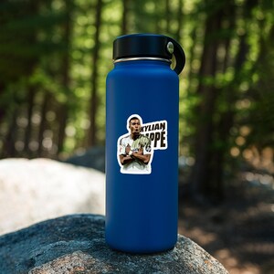 May include: A blue insulated water bottle with a black lid and handle. A sticker of Kylian Mbappe in a white jersey is affixed to the bottle. The sticker has the name "KYLIAN MBAPPE" in white and black.