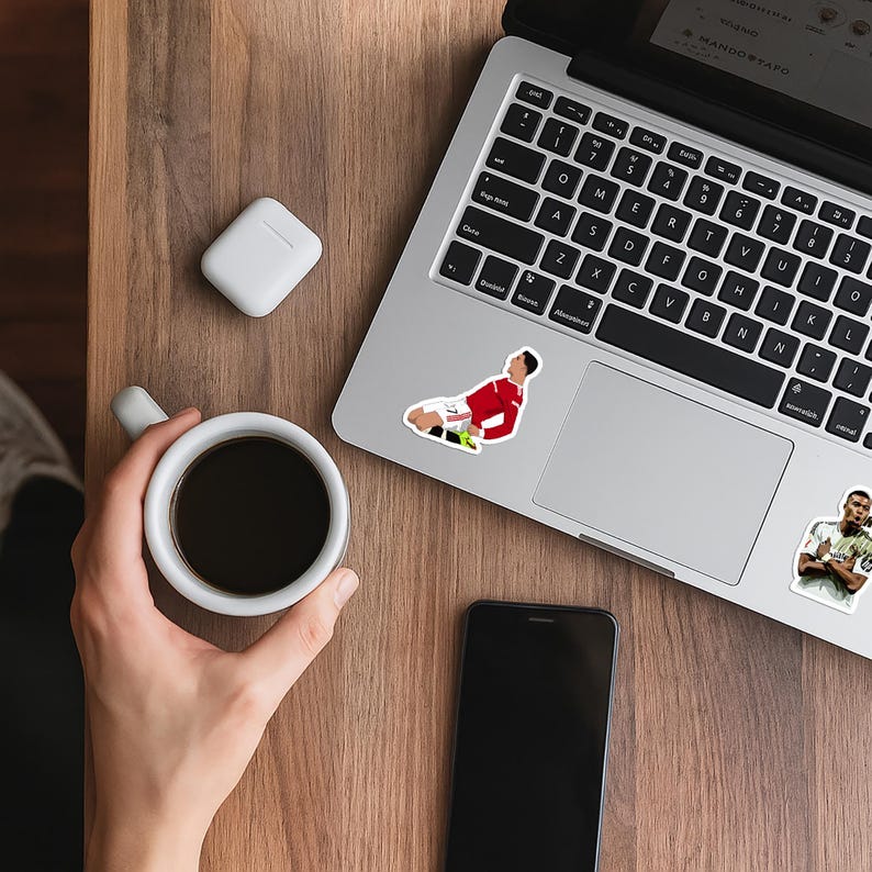 May include: A laptop with stickers of soccer players, a smartphone, a white wireless earbud case, and a cup of coffee on a wooden surface. The laptop keyboard is visible. A hand holds the coffee cup. The laptop screen displays text.