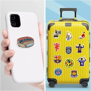 May include: A white phone case with a sticker of a stadium and a yellow suitcase covered in soccer team stickers. The suitcase has a retractable handle and wheels. Stickers include team logos and player images.