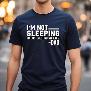 May include: Navy blue t-shirt with white text that reads "I'M NOT SLEEPING I'M JUST RESTING MY EYES -DAD". The shirt is a short-sleeved crew neck style.