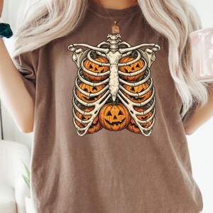 May include: A brown short-sleeve t-shirt featuring a graphic print of a human rib cage. Inside the rib cage, there are five orange jack-o'-lantern pumpkins with carved faces. The design is centered on the chest of the garment.