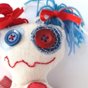 May include: A handmade fabric doll with a heart-shaped body and stitched features. The doll has blue and red accents, including button eyes, a stitched smile, and yarn hair tied with red ribbons. The doll is a whimsical, handcrafted item.