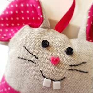 Puede incluir: Decoración de conejo de Pascua hecha a mano con una cara de lino gris, ojos de botón negros y una nariz rosa en forma de corazón. El conejo tiene orejas rosas con lunares y una cinta a juego. El texto "crumb, joie & ama" es visible.