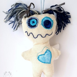Könnte beinhalten: Handgemachte Puppe mit einem skurrilen Design. Die Puppe hat einen cremefarbenen Stoffkörper, blaue Knopfaugen und einen gestickten Mund. Sie hat blaue und graue Garnhaare und ein blaues Herz auf der Brust. Kleine Engelsflügel sind angebracht.
