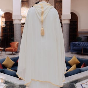 May include: A cream-colored hooded robe with gold trim and a matching tassel. The robe is worn by a person standing in a courtyard with pillars and seating. The robe appears to be made of a lightweight fabric.