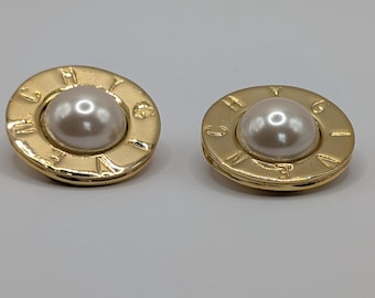 May include: A pair of gold-coloured clip-on earrings. Each earring has a large, faux pearl centre surrounded by a gold-coloured frame. The frame has embossed lettering. The earrings are set against a plain, light background.
